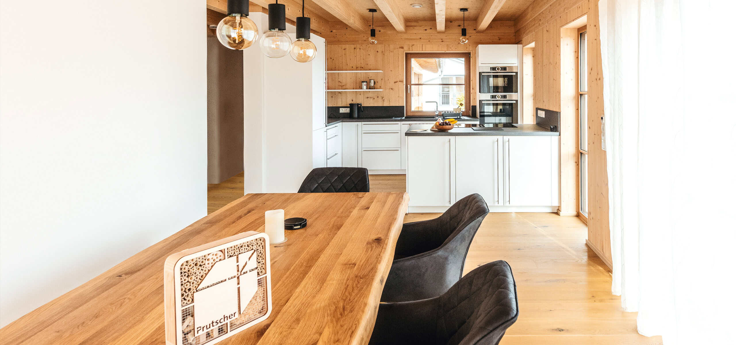 Moderne Küche mit Essbereich, deren Wände, Decke und Boden aus Holz sind. Im Vordergrund steht ein Holztisch mit schwarzen Stühlen, im Hintergrund sind weiße Schränke und Einbaugeräte zu sehen. Große Fenster lassen Tageslicht herein.