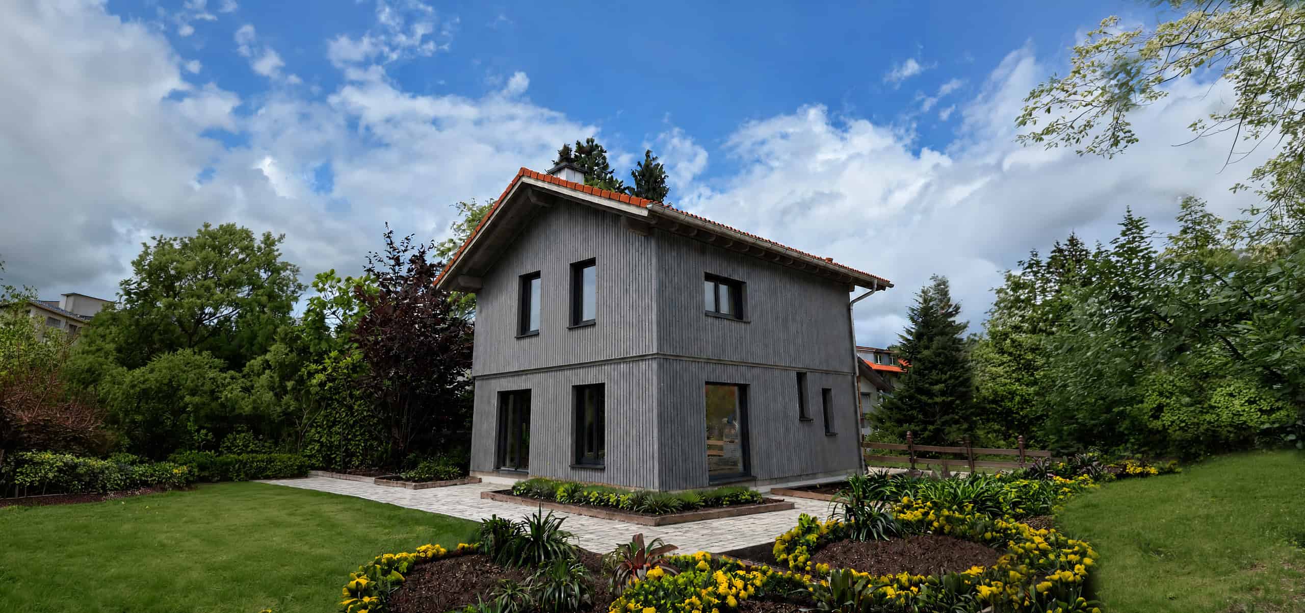 Ein modernes, graues, zweistöckiges Haus mit rotem Ziegeldach ist von einem gepflegten Garten mit gelben Blumen, grünen Sträuchern und Bäumen umgeben, über dem sich ein teilweise bewölkter blauer Himmel erstreckt.