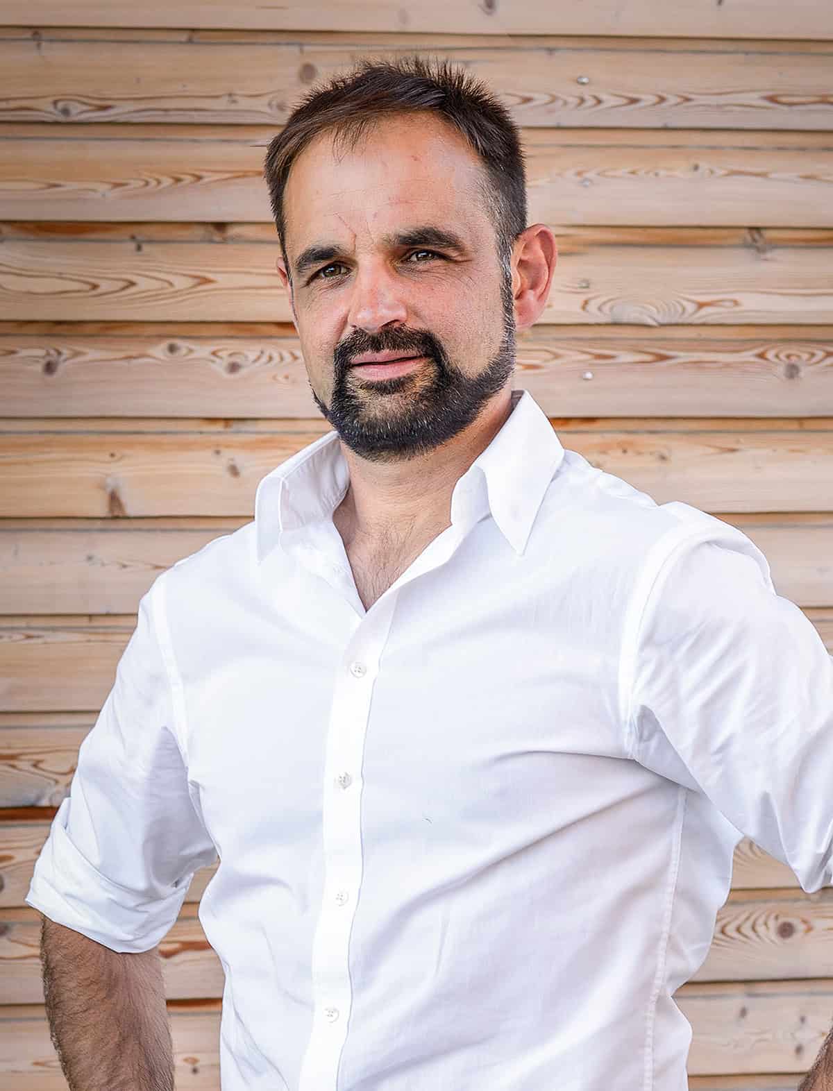 Ein Mann mit gepflegtem Bart und Schnurrbart, der ein weißes Hemd trägt, steht vor einer Holzwand und blickt mit einem leichten Lächeln in die Kamera.