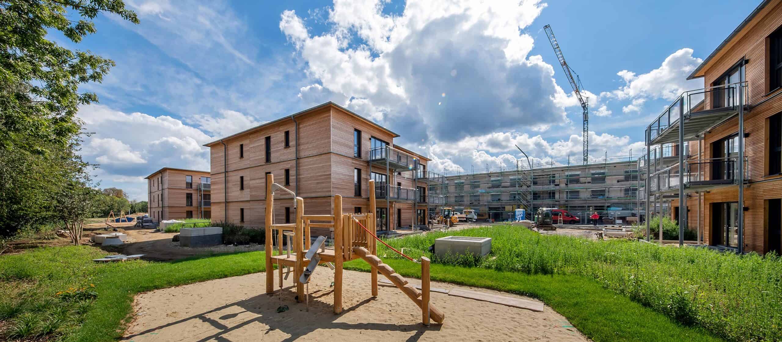 Ein moderner Spielplatz mit Spielgeräten aus Holz befindet sich inmitten neuer Wohnhäuser aus Holz; im Hintergrund sind unter einem teilweise bewölkten Himmel.