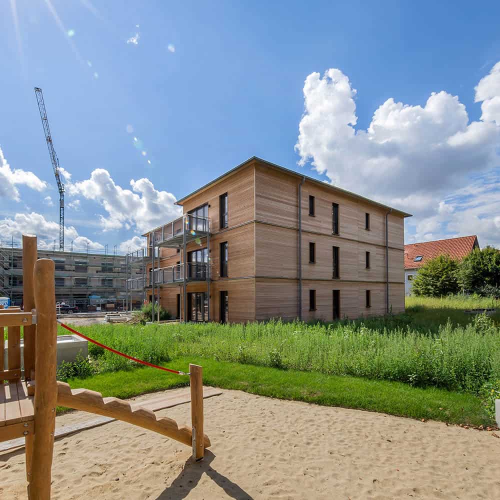 Unter einem blauen Himmel mit vereinzelten Wolken steht ein modernes Mehrfamilienhaus aus Holz in der Nähe einer Wiese und eines Sandspielplatzes. Im Hintergrund sind weitere Gebäude zu sehen.