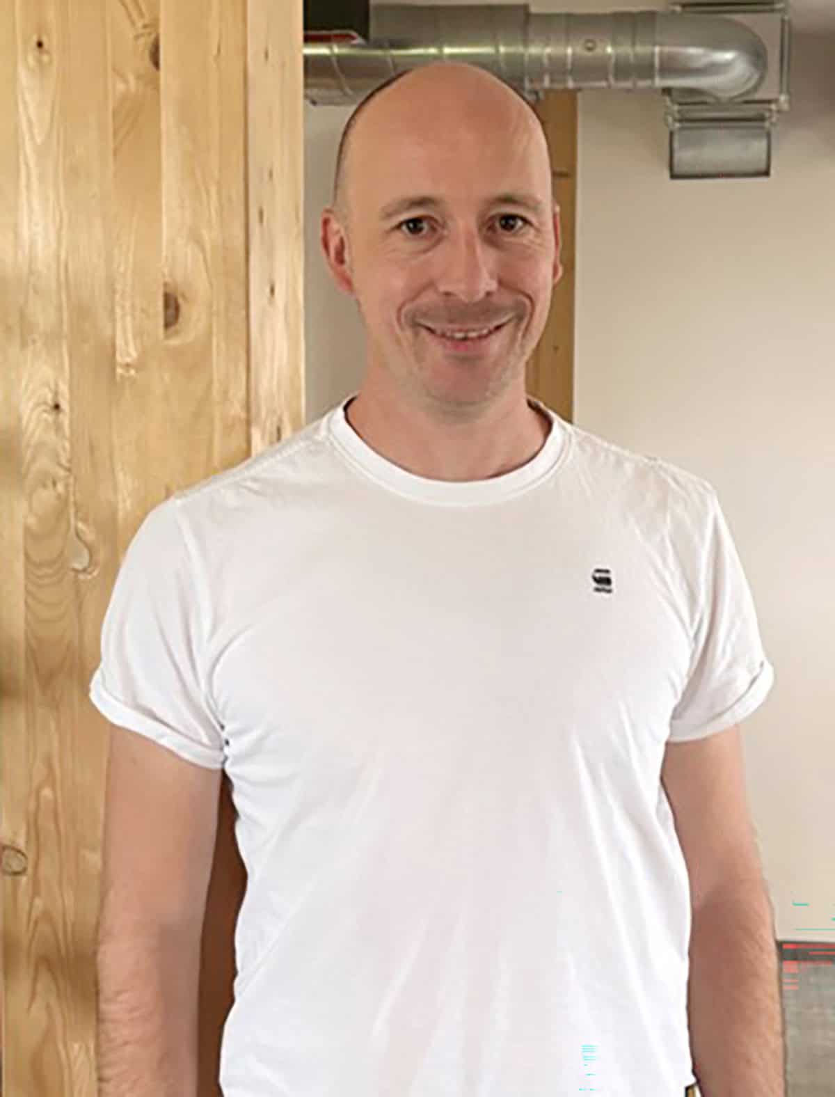 Ein lächelnder Mann in einem weißen T-Shirt steht in einem Raum neben einer Holzwand.