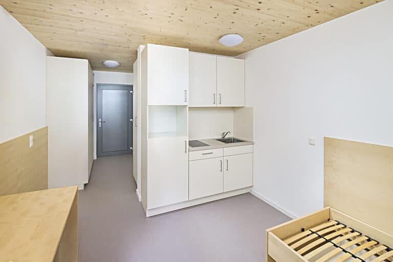 Minimalistisch eingerichtetes Wohnheimzimmer mit einer kleinen Kochnische, eingebauten Holzschränken und einem leeren Bettgestell – ideal als schlüsselfertiger Wohnraum für Mitarbeiter. Die helle Holzdecke und die weißen Wände sorgen für einen klaren, modernen Look.