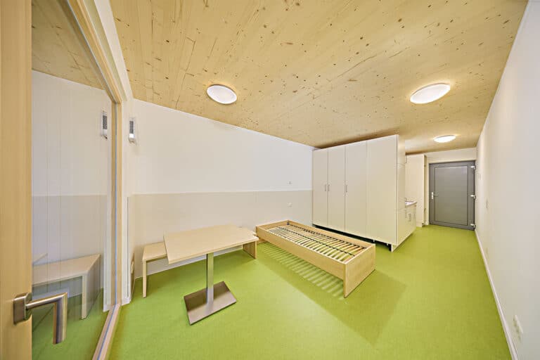 Ein minimalistischer Raum mit grünem Bodenbelag, einem Bettgestell, einem kleinen Tisch und einem Stuhl, zwei weißen Kleiderschränken, einem Waschbecken und einer Tür. Eine Decke aus hellem Holz und eine Glastrennwand schaffen einen schlüsselfertigen Wohnraum.