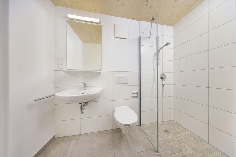 Minimalistisches Badezimmer mit weiß gefliesten Wänden, einem wandhängenden Waschbecken, einem wandhängenden WC und einer begehbaren Dusche mit Glastrennwand – perfekt für schlüsselfertigen Wohnraum im modernen Design.