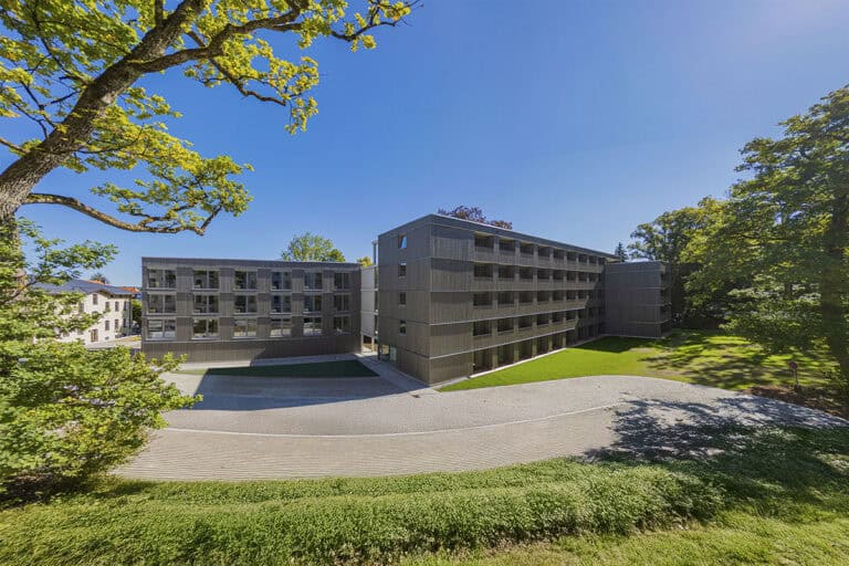 Moderne, dreistöckige, graue Mehrfamilienhäuser mit Balkonen bieten schlüsselfertigen Wohnraum zum Festpreis, umgeben von grünem Rasen, Bäumen und einem strahlend blauen Himmel an sonnigen Tagen.