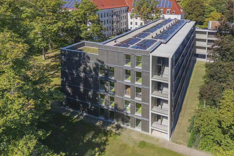 Ein modernes Mehrfamilienhaus mit Balkonen und Solaranlagen auf dem Dach bietet schlüsselfertigen Wohnraum, umgeben von Bäumen und Grünflächen. Im Hintergrund sind weitere Festpreis-Gebäude mit Solaranlagen zu sehen.