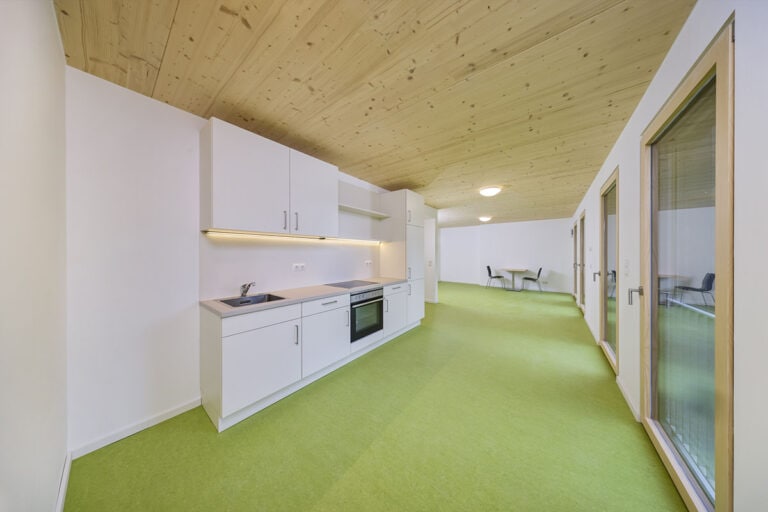 Moderne Küche mit weißen Schränken und Einbaugeräten auf der linken Seite, grünem Bodenbelag, Holzdecke und großen Fenstern – ein schlüsselfertiger Wohnraum für Mitarbeiter mit einem gemütlichen Essbereich am Ende des Raums.