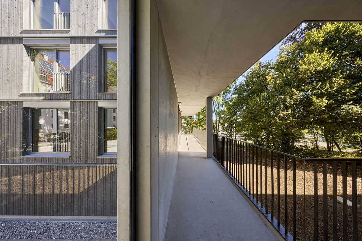 Blick auf ein modernes Wohnhaus mit Holzfassade auf der linken Seite und einem langen, offenen Gang mit Metallgeländern, von dem aus man auf grüne Bäume blickt – idealer schlüsselfertiger Wohnraum für Mitarbeiter.