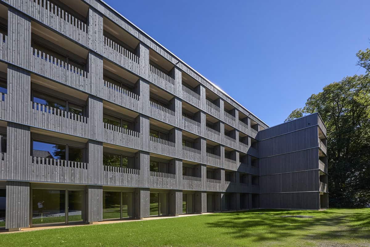 Modernes vierstöckiges Gebäude mit Holzvertäfelung, großen Fenstern und Balkonen. Dieser schlüsselfertige Wohnraum liegt auf einer Rasenfläche unter strahlend blauem Himmel, im Hintergrund sind Bäume zu sehen.